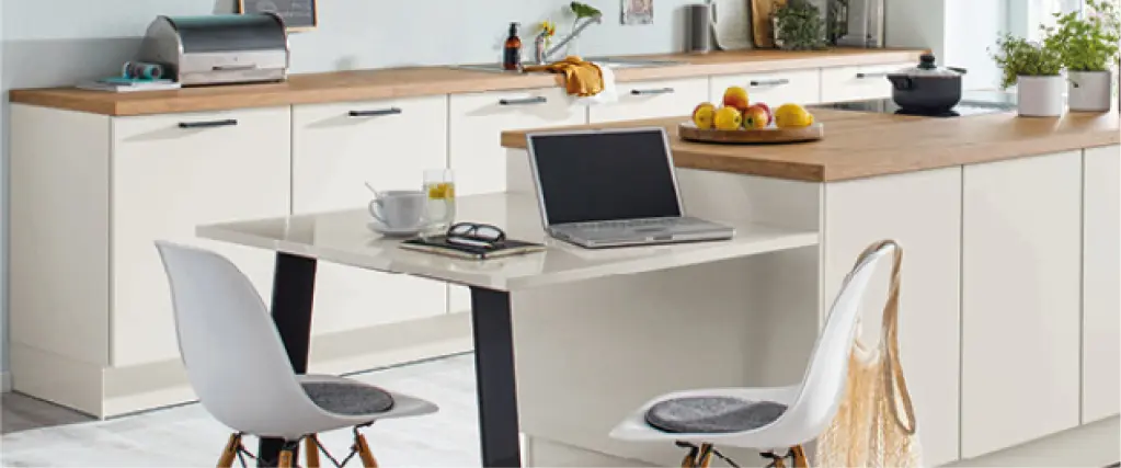 Helle Küche mit Kochinsel, Holzarbeitsplatte und integriertem Essplatz. Moderner Küchentisch mit Stühlen, Laptop und Geschirr auf der Arbeitsfläche. Funktionale Küchenplanung für Wohnen und Arbeiten im Alltag.